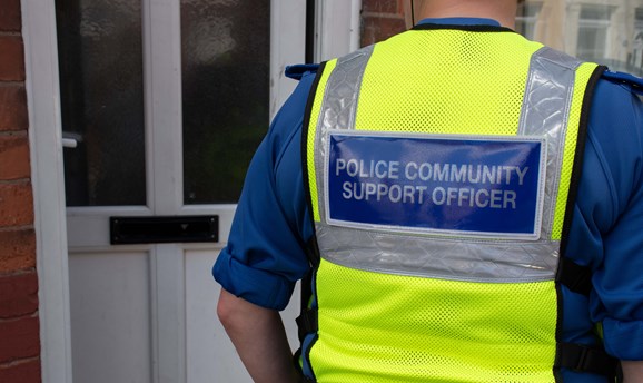 Image of the back of a police community support officers vest stood at and door. 